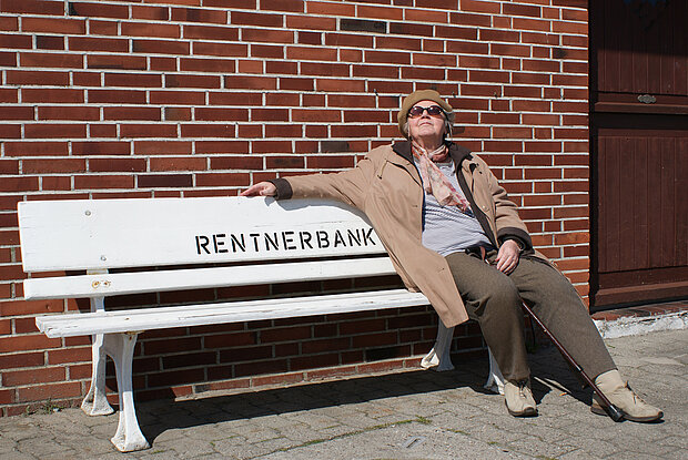 ältere Dame sitzt auf einer Bank im Freien mit dem Schriftzug "Rentnerbank"