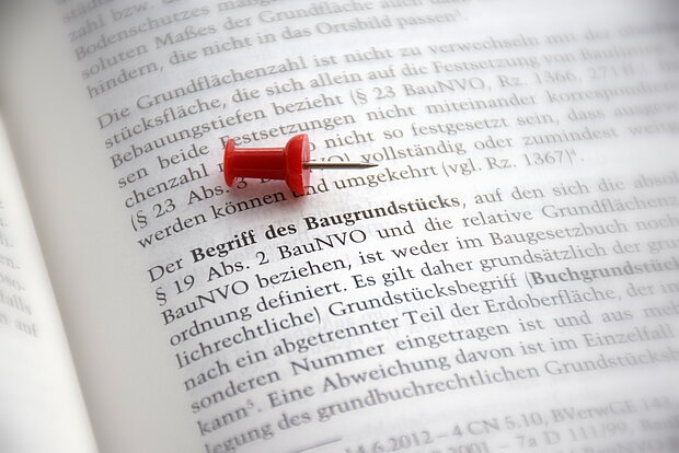 Reißnadel liegt auf einem aufgeschlagenen Buch bei dem Begriff "Begriff des Baugrundstücks"